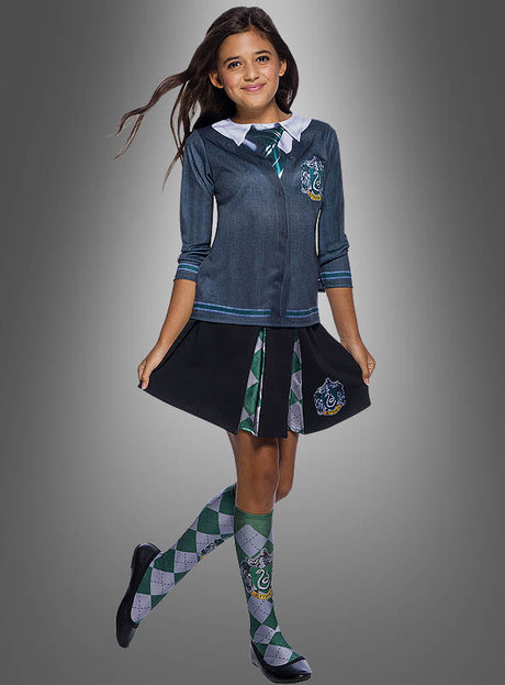 Ein Mädchen, das das Slytherin-Shirt aus „Harry Potter“ für Kinder trägt – dazu eine grüne Krawatte, einen Faltenrock und karierte Socken –, steht vor einem grauen Hintergrund.