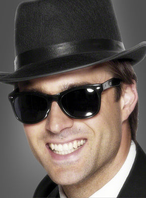 Ein Mann mit schwarzem Fedora-Hut, schwarzem Anzug und einer Sonnenbrille im Stil der 50er Jahre lächelt vor einem schlichten grauen Hintergrund – ein Look, der an das klassische Blues-Brothers-Kostüm erinnert.