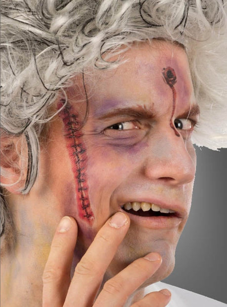 Ein Mann mit weißem, zerzaustem Haar und theatralischem Make-up – darunter eine genähte Narbe auf der Wange und eine künstliche Wunde auf der Stirn, die mit „Blutige Narben Tattoo Wunden“ gestaltet wurde – berührt mit besorgtem Gesichtsausdruck sein Gesicht.