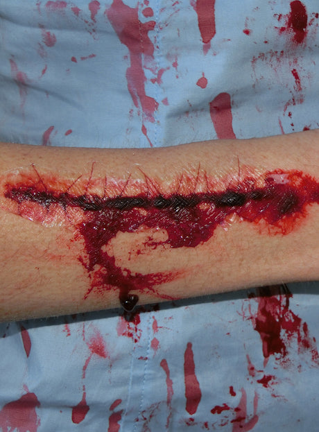 Ein Unterarm mit einer großen, genähten Wunde, umgeben von Blut und Blutspritzern auf blauem Stoffhintergrund – dieses eindrucksvolle Beispiel für die „Blutige Narben Tattoo Wunden“ eignet sich perfekt für Halloween-Narben-Tattoos und besticht durch realistisches Kunstblut für einen besonders gruseligen Effekt.