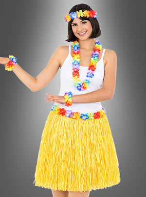 Die Person trägt das 5-teilige Hawaii-Set mit Rock und Accessoires – bestehend aus einem gelben Grasrock, einem weißen Trägertop und buntem Blumenschmuck an Kopf, Hals und Handgelenken – und steht vor einem grauen Hintergrund.