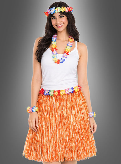 Eine Frau steht lächelnd vor einem grauen Hintergrund und trägt ein weißes Trägertop sowie das 5-teilige Hawaii-Set mit Rock und Accessoires, zu dem ein orangefarbener Grasrock und bunte Blumen-Accessoires gehören.