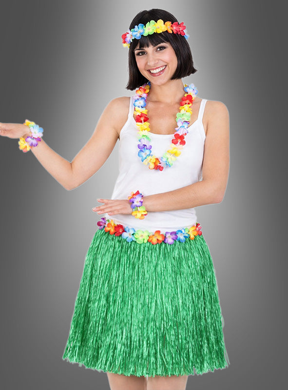 Eine Frau, die das 5-teilige Hawaii-Set mit Rock und Accessoires trägt – bestehend aus einem grünen Grasrock (Hawaii-Rock), einem weißen Trägertop und bunten Blumenaccessoires (Blumenschmuck) –, posiert lächelnd vor einem grauen Hintergrund und präsentiert damit den perfekten festlichen, tropischen Look.