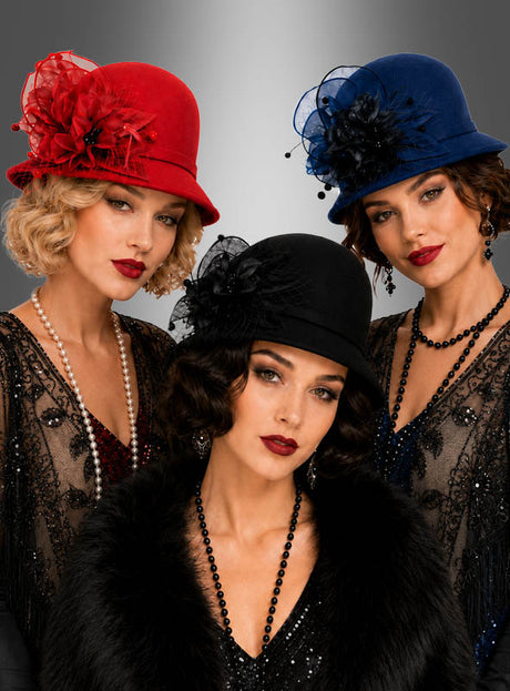 Drei Frauen in Kleidern und Hüten im Vintage-Stil in Rot, Blau und Schwarz, die jeweils mit Perlenketten geschmückt sind – darunter ein auffälliger hochwertiger Glockenhut aus Filz im Stil der 20er Jahre als Gatsby-Accessoire –, posieren elegant vor einem grauen Hintergrund mit Farbverlauf.