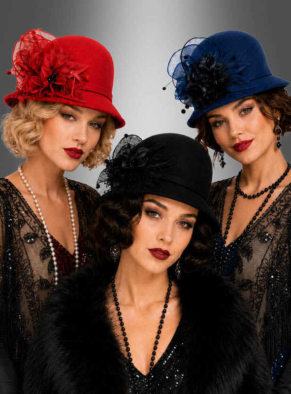 Drei Frauen in Kleidern und Hüten im Vintage-Stil in Rot, Blau und Schwarz, die jeweils mit Perlenketten geschmückt sind – darunter ein auffälliger hochwertiger Glockenhut aus Filz im Stil der 20er Jahre als Gatsby-Accessoire –, posieren elegant vor einem grauen Hintergrund mit Farbverlauf.