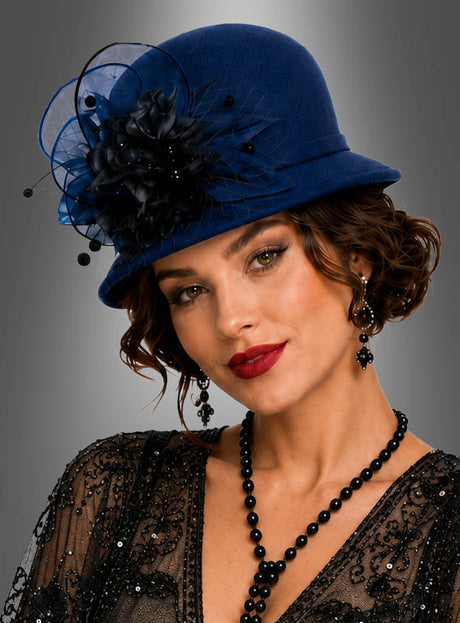 Eine Frau, die einen hochwertigen Glockenhut aus Filz im Stil der 20er Jahre in Blau mit schwarzer Blumenverzierung trägt, ergänzt durch eine schwarze Perlenkette und ein schwarzes Spitzenkleid, steht mit dem Blick nach vorne vor einem neutralen grauen Hintergrund – ein ikonisches Gatsby-Accessoire, das die Eleganz der 20er Jahre perfekt verkörpert.
