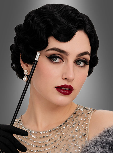 Frau mit kurzem, welligem schwarzem Haar, frisiert im Stil der „20er Wasserwelle Premium Perücke Barbara“, in einem mit Perlen bestickten Kleid, mit dunklem Make-up, Perlenohrring und schwarzen Handschuhen, die vor einem neutralen Hintergrund eine Zigarettenspitze hält.