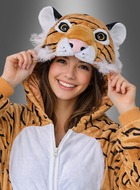 Eine lächelnde Frau trägt das „Deluxe Tiger Kostüm aus Plüsch“ mit einer Kapuze in Form eines Tigergesichts und hält die Ränder der Kapuze mit beiden Händen vor einem grauen Hintergrund mit Farbverlauf fest.