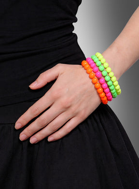 Eine schwarz gekleidete Person trägt das „Neon Perlenarmbänder 4er Set 80er“ – vier farbenfrohe Perlenarmbänder in Orange, Pink, Gelb und Grün – am Handgelenk, während sie die Hand in die Hüfte stützt. Diese auffälligen Armbänder sind das perfekte Accessoire, um jedes retro-inspirierte Outfit abzurunden.