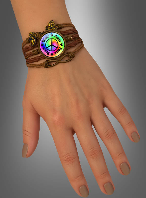 Eine Hand, an der das „Peace Armband“ in Braun getragen wird – ein geflochtenes Armband mit einem runden Anhänger, auf dem ein Regenbogen-Friedenszeichen und die Worte „Peace & Love“ zu sehen sind –, was es zu einem idealen Festival-Accessoire oder Hippie-Schmuckstück macht.