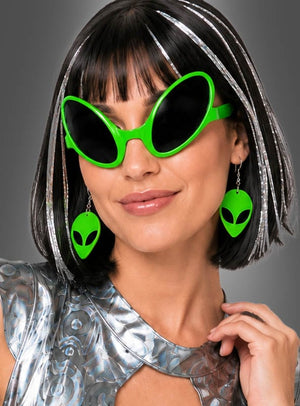 Eine Frau mit einer schwarzen Bob-Perücke mit silbernen Strähnchen trägt eine grüne Sonnenbrille in Alien-Form und kombiniert diese mit Alien-Ohrringen für Weltraumkostüme. Ihren Look rundet ein glänzendes silbernes Weltraumkostüm ab, das vor einem grauen Hintergrund zur Geltung kommt.