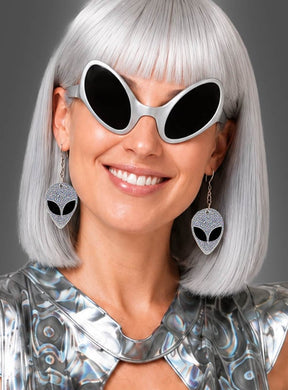 Eine Frau mit silbernem Haar, die eine große schwarze Sonnenbrille und ein metallisches Oberteil trägt, lächelt vor einem schlichten grauen Hintergrund, während sie die „Alien-Ohrringe für Space-Kostüme“ präsentiert – Ohrringe in Form von silbernen Aliens.