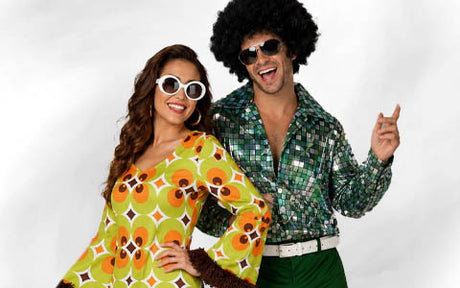 Zwei Personen in Retro-Outfits aus den 1970er Jahren, die Sonnenbrillen tragen und lächeln; die eine trägt ein gemustertes Kleid, die andere ein glänzendes Hemd und eine Afro-Perücke.