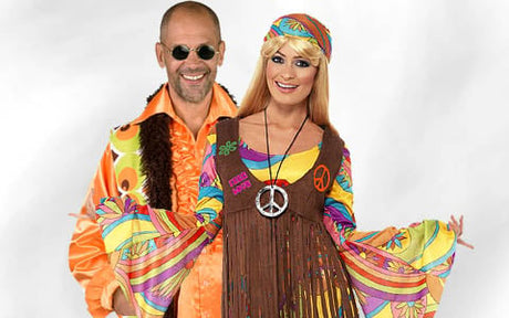 Zwei Erwachsene in bunten, von den 1960er Jahren inspirierten Hippie-Kostümen: Der eine trägt eine runde Sonnenbrille und ein orangefarbenes Hemd, der andere ein Stirnband, eine Weste mit Fransen und eine Halskette mit Peace-Zeichen; beide lächeln.
