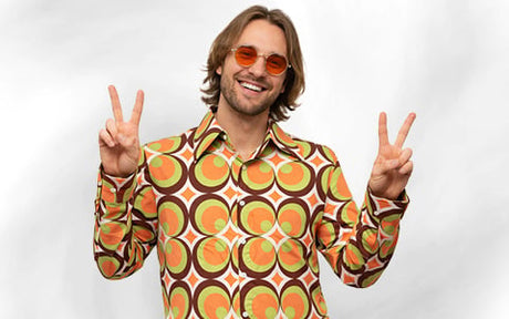 Ein lächelnder Mann in einem Hemd mit Retro-Muster und einer orangefarbenen Brille streckt vor einem schlichten weißen Hintergrund beide Hände mit Peace-Zeichen in die Höhe.