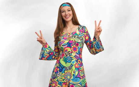 Eine Frau in einem bunten Kleid im Stil der 1960er Jahre und mit einem Stirnband formt mit beiden Händen das Peace-Zeichen und lächelt in die Kamera.