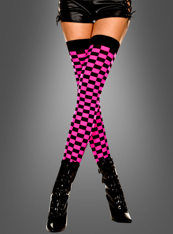 Eine Person, die schwarze Shorts, schwarze Stiefel mit hohen Absätzen sowie rosa-schwarze, halterlose „Schachbrett“-Overknees im auffälligen Ska-Look trägt, steht mit gekreuzten Beinen vor einem grauen Hintergrund.