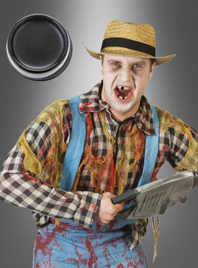 Eine Person mit Strohhut und zerlumpter Kleidung, deren Zähne mit Zahnschwarz-Make-up schwarz gefärbt sind und bei der einige Zähne fehlen, steht mit einem Werkzeug in der Hand vor einem grauen Hintergrund, in dessen Ecke sich ein großer schwarzer Knopf befindet.