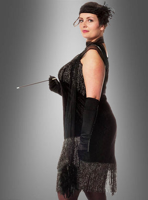 Eine Frau in der Flapper-Mode der 1920er Jahre mit einem schwarzen XXL-Fransenkleid „Diva 20er Jahre“, eleganten Handschuhen, einem Stirnband und einer Zigarettenspitze, die im Profil vor einem grauen Hintergrund steht.