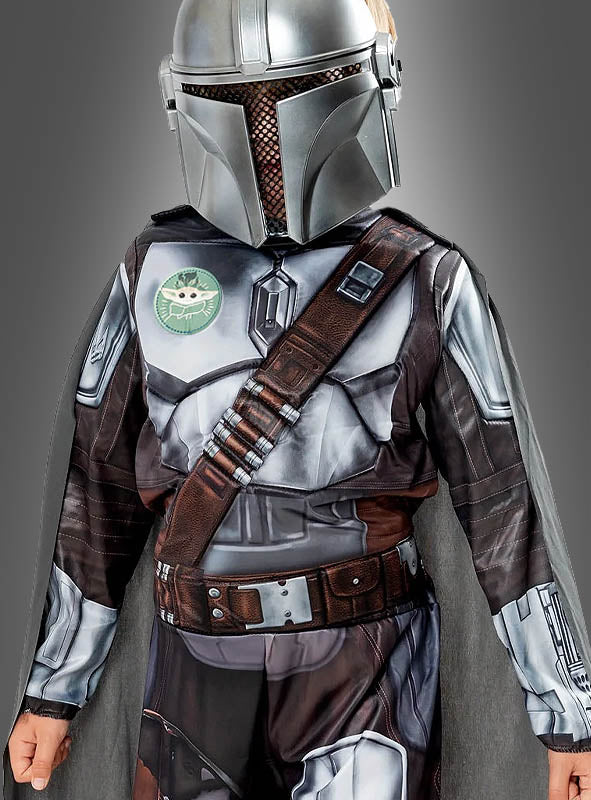 Ein Kind im Mandalorianer-Kinderkostüm aus „Star Wars“, komplett mit Helm, Rüstungsdetails, Grogu-Emblem und braunem Patronengurt, steht vor einem grauen Hintergrund – ideal für jeden jungen Fan, der ein authentisches „Star Wars“-Kostüm sucht.