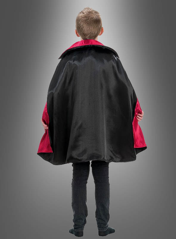 Ein Kind steht mit dem Rücken zur Kamera und trägt das Vampir-Kostüm „Theodor“ – einen schwarzen Umhang mit auffälligem rotem Futter –, das sich perfekt für jedes gruselige Halloween-Kostüm oder als klassisches Vampir-Kostüm für Kinder eignet. Im Hintergrund ist ein grauer Farbverlauf zu sehen.