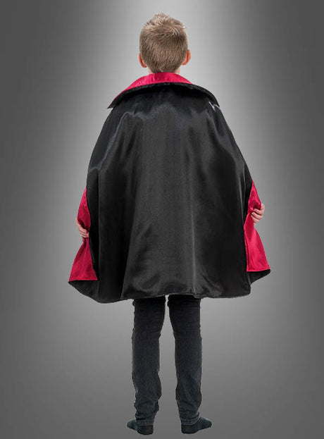 Ein Kind steht mit dem Rücken zur Kamera und trägt das Vampir-Kostüm „Theodor“ – einen schwarzen Umhang mit auffälligem rotem Futter –, das sich perfekt für jedes gruselige Halloween-Kostüm oder als klassisches Vampir-Kostüm für Kinder eignet. Im Hintergrund ist ein grauer Farbverlauf zu sehen.