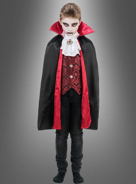 Ein Kind im Vampir-Kostüm „Theodor“ – mit einem auffälligen rot-schwarzen Umhang, einem Rüschenhemd und Make-up – steht selbstbewusst vor einem grauen Hintergrund.