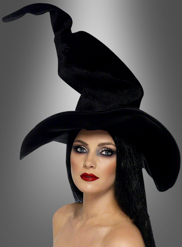 Eine Frau mit langen schwarzen Haaren und auffälligem Make-up trägt den „Klassischen Hexenhut schwarz“ – ein markantes Accessoire für Hexenkostüme – vor einem grau verlaufenden Hintergrund.