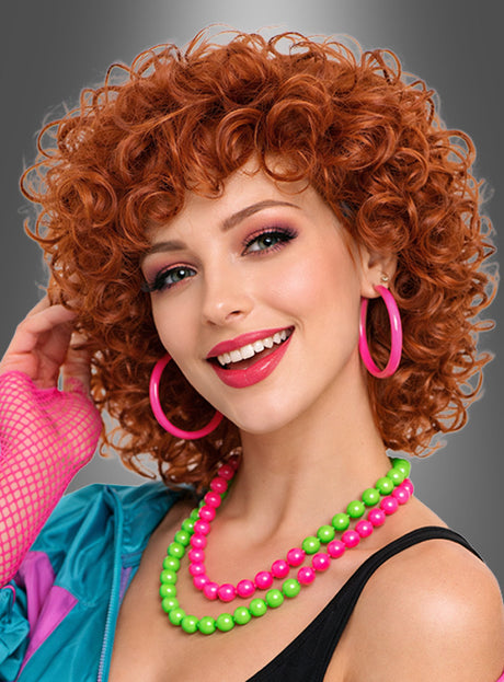 Eine lächelnde Frau trägt die Perücke „Halblange Locken Perücke Dauerwelle Cindy“ mit roten Locken, dazu rosa Creolen, auffälliges Make-up sowie neonrosa und neongrüne Perlenketten; sie ist in einem Retro-Outfit gekleidet und steht vor einem grauen Hintergrund.