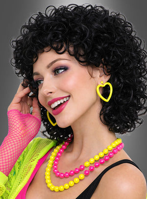 Eine Frau mit kurzen, lockigen schwarzen Haaren, die als Perücke „Halblange Locken Perücke Dauerwelle Cindy“ gestylt sind, trägt leuchtend gelbe Herzohrringe, rosa und gelbe Perlenketten, einen rosa Netzhandschuh und ein neongrünes Oberteil und lächelt vor einem grauen Hintergrund.