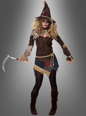 Eine Person im Vogelscheuche-Kostüm mit Sense, bestehend aus einem spitzen Hut, einem Patchwork-Kleid und einer Sense in der Hand, steht vor einem schlichten grauen Hintergrund – ein ideales Halloween-Kostüm für alle, die auf der Suche nach einem gruseligen Kostüm sind.