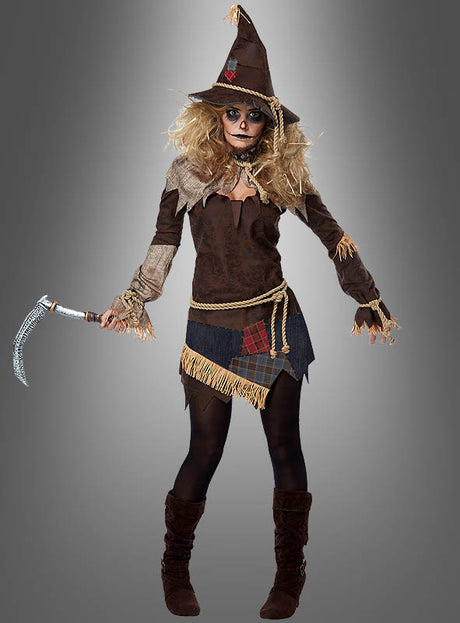 Eine Person im Vogelscheuche-Kostüm mit Sense, bestehend aus einem spitzen Hut, einem Patchwork-Kleid und einer Sense in der Hand, steht vor einem schlichten grauen Hintergrund – ein ideales Halloween-Kostüm für alle, die auf der Suche nach einem gruseligen Kostüm sind.