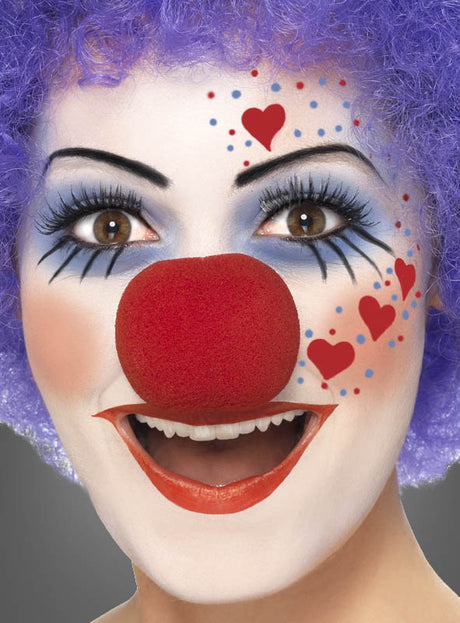 Nahaufnahme einer Person mit Clown-Schminke, die mit der „Schminke Farbpalette Fasching für Kinder und Erwachsene“ aufgetragen wurde: rote Nase, lila Perücke und auf die Wange gemalte Herzen; die Person lächelt in die Kamera.