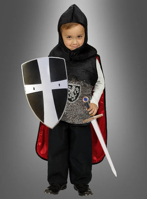 Ein kleines Kind in einem Ritterkostüm hält das „Kinder Ritter Schild mit Schwert 48 cm“ in den Händen, das mit einem schwarz-weißen Kreuz verziert ist und den Geist von „Tempelritter Spielzeug“ verkörpert. Das Kind steht stolz vor einem schlichten grauen Hintergrund.