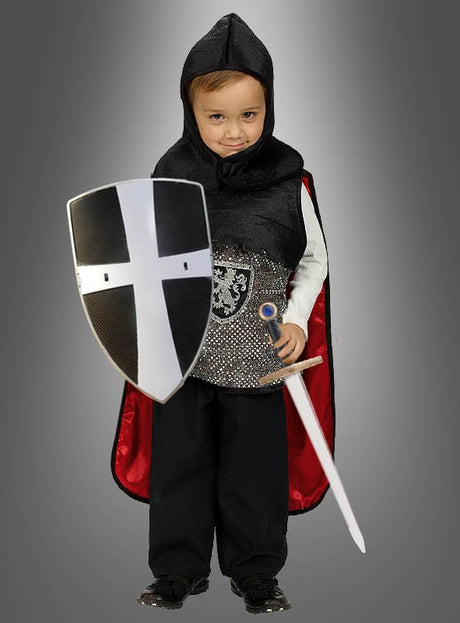 Ein kleines Kind in einem Ritterkostüm hält das „Kinder Ritter Schild mit Schwert 48 cm“ in den Händen, das mit einem schwarz-weißen Kreuz verziert ist und den Geist von „Tempelritter Spielzeug“ verkörpert. Das Kind steht stolz vor einem schlichten grauen Hintergrund.
