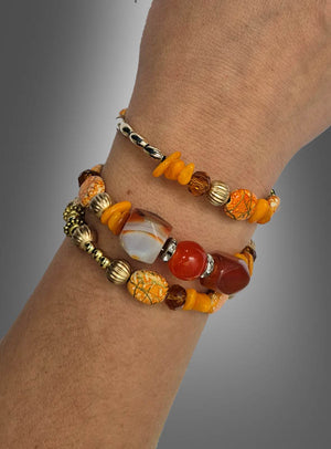 Mit dem Armreif „Perlen“ im Hippie-Stil sorgen zwei Perlenarmbänder in leuchtendem Orange, Braun, Gelb und Metallic-Tönen – akzentuiert durch eine auffällige marmorierte Perle und strukturierte orangefarbene Steine – vor einem grauen Hintergrund mit Farbverlauf für den perfekten Boho-Look.