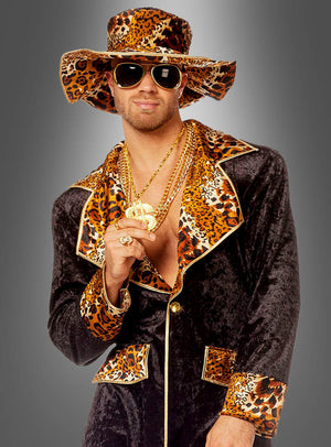Vor einem grauen Hintergrund steht ein Mann, der das „Cooles Pimp Outfit Leopardenmuster“ trägt – bestehend aus einem Hut mit Leopardenmuster, einer Sonnenbrille, Goldschmuck und einem schwarzen Samtanzug mit passenden Akzenten im Leopardenmuster –, was ihm einen auffälligen Look verleiht.