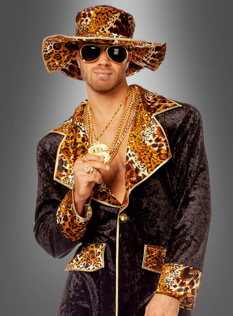 Vor einem grauen Hintergrund steht ein Mann, der das „Cooles Pimp Outfit Leopardenmuster“ trägt – bestehend aus einem Hut mit Leopardenmuster, einer Sonnenbrille, Goldschmuck und einem schwarzen Samtanzug mit passenden Akzenten im Leopardenmuster –, was ihm einen auffälligen Look verleiht.