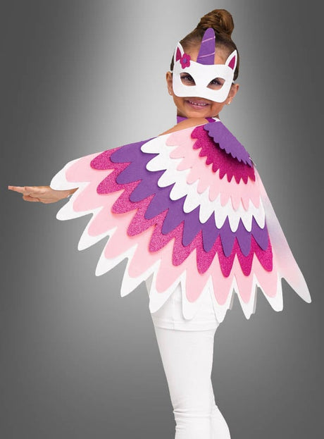 Ein Kind in einem Einhorn-Kostüm, komplett mit passender Maske und einem Glitzer-Umhang mit Flügeln für Kinder, der aus bunten, rosa, lila und weißen, federartigen Lagen besteht, steht vor einem grauen Hintergrund.