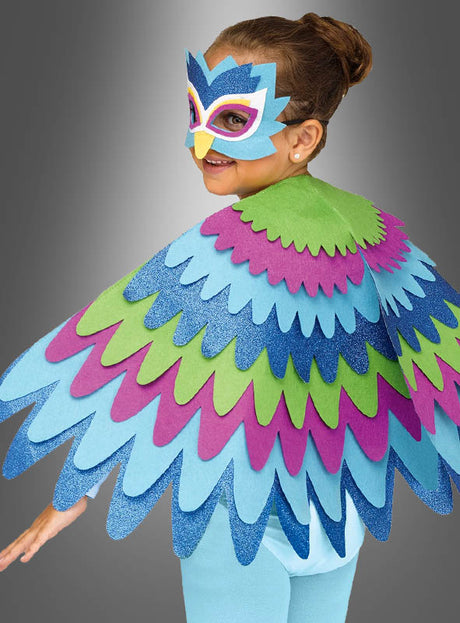 Ein Kind in einem bunten Vogelkostüm mit einem mehrlagigen, federförmigen „Glitzer Cape Flügel für Kinder“ und einer passenden Vogelmaske blickt vor einem grauen Hintergrund über die Schulter.