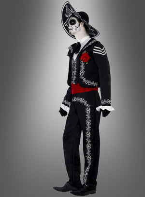 Eine Person, die das „Tag der Toten“-Bräutigam-Kostüm trägt – bestehend aus einer von Mariachi inspirierten Skelett-Gesichtsbemalung, einem schwarzen Hut und einem Anzug, der mit weißer Stickerei, einer roten Schärpe und einer Rose verziert ist –, steht vor einem grauen Hintergrund mit Farbverlauf.