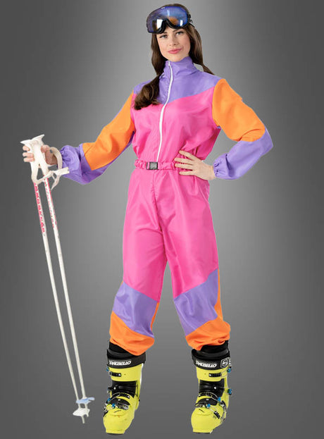 Eine Frau im rosa „Cooler Skianzug 80er Design“ – mit leuchtend orangefarbenen und violetten Einsätzen – hält weiße Skistöcke in der Hand, trägt gelbe Skischuhe und eine Skibrille und steht vor einem grauen Hintergrund. Damit eignet sich dieser Retro-Look ideal für eine Après-Ski-Party oder als auffälliges 80er-Jahre-Kostüm.