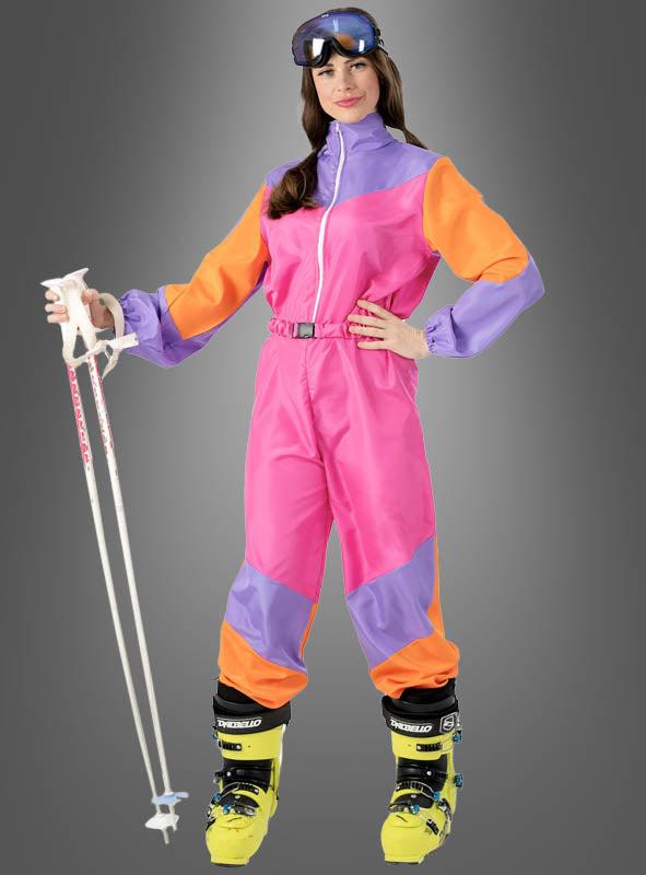 Eine Frau im rosa „Cooler Skianzug 80er Design“ – mit leuchtend orangefarbenen und violetten Einsätzen – hält weiße Skistöcke in der Hand, trägt gelbe Skischuhe und eine Skibrille und steht vor einem grauen Hintergrund. Damit eignet sich dieser Retro-Look ideal für eine Après-Ski-Party oder als auffälliges 80er-Jahre-Kostüm.
