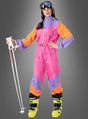 Eine Frau im rosa „Cooler Skianzug 80er Design“ – mit leuchtend orangefarbenen und violetten Einsätzen – hält weiße Skistöcke in der Hand, trägt gelbe Skischuhe und eine Skibrille und steht vor einem grauen Hintergrund. Damit eignet sich dieser Retro-Look ideal für eine Après-Ski-Party oder als auffälliges 80er-Jahre-Kostüm.