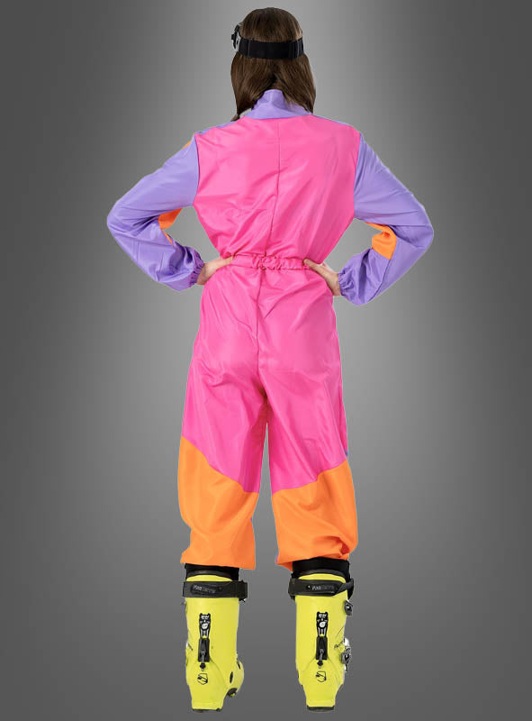 Eine Person steht mit dem Rücken zur Kamera und trägt den „Cooler Skianzug 80er Design“ in Pink – einen farbenfrohen, von den 80er Jahren inspirierten Skianzug in Rosa-, Lila- und Orangetönen –, dazu gelbe Skischuhe, vor einem grauen Hintergrund mit Farbverlauf; perfekt für jede Après-Ski-Party.