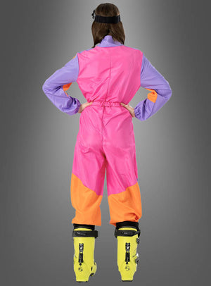 Eine Person steht mit dem Rücken zur Kamera und trägt den „Cooler Skianzug 80er Design“ in Pink – einen farbenfrohen, von den 80er Jahren inspirierten Skianzug in Rosa-, Lila- und Orangetönen –, dazu gelbe Skischuhe, vor einem grauen Hintergrund mit Farbverlauf; perfekt für jede Après-Ski-Party.