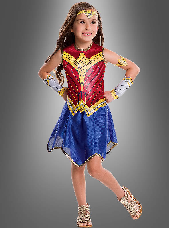 Vor einem schlichten grauen Hintergrund steht ein junges Mädchen lächelnd, gekleidet in ein farbenfrohes Wonder-Woman-Kinderkostüm aus der „Justice League“-Reihe, bestehend aus einem rot-gold-blauen Outfit, goldenen Sandalen und einem Stirnband.