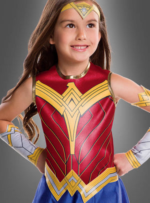 Ein junges Mädchen lächelt selbstbewusst in ihrem Wonder Woman-Kinderkostüm aus der Justice League mit leuchtenden goldenen, roten und blauen Details, während sie mit den Händen in die Hüften gestemmt vor einem schlichten grauen Hintergrund steht. Dieses bezaubernde DC-Comics-Kostüm ist perfekt für jede angehende Superheldin!.
