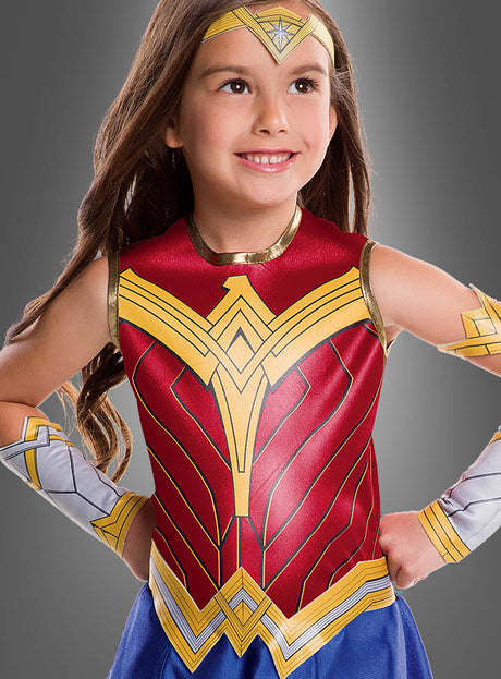 Ein junges Mädchen lächelt selbstbewusst in ihrem Wonder Woman-Kinderkostüm aus der Justice League mit leuchtenden goldenen, roten und blauen Details, während sie mit den Händen in die Hüften gestemmt vor einem schlichten grauen Hintergrund steht. Dieses bezaubernde DC-Comics-Kostüm ist perfekt für jede angehende Superheldin!.