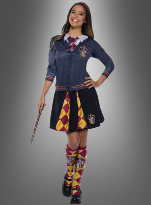 Eine Person in einem Kostüm im Gryffindor-Stil hält einen Zauberstab in der Hand und trägt das bedruckte Harry-Potter-Shirt „Gryffindor“ sowie eine Strickjacke, eine Krawatte, einen Rock und hohe Socken mit gelben und roten Mustern auf grauem Hintergrund – perfekt für jeden Harry-Potter-Kostüm-Fan.